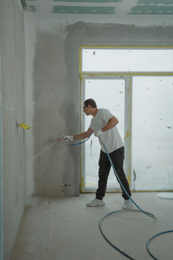 A man uses a spray gun to paint an interior wall in a room under renovation.